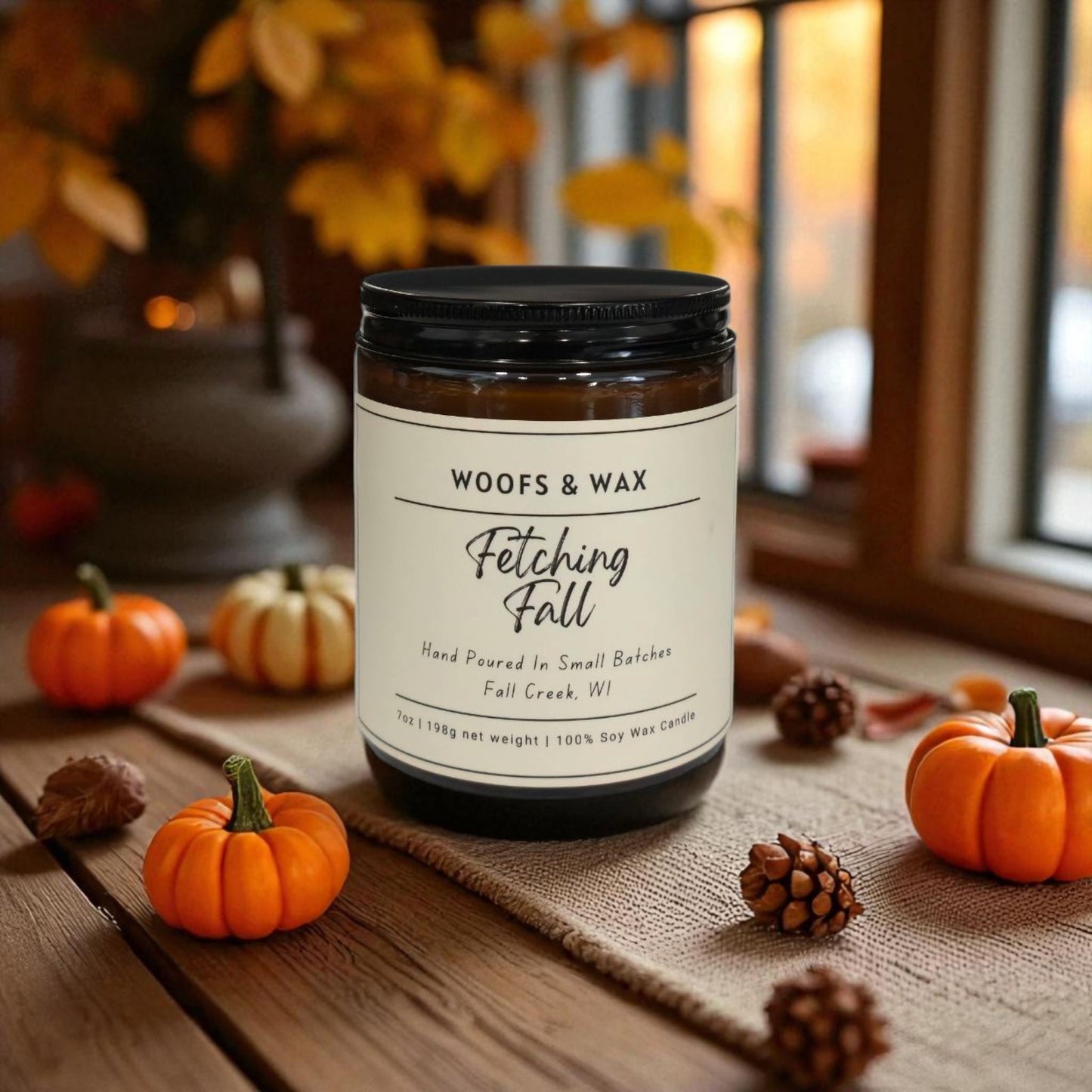 Candle labeled 'Woofs & Wax Fetching Fall' on a wooden surface with pumpkins and pinecones.