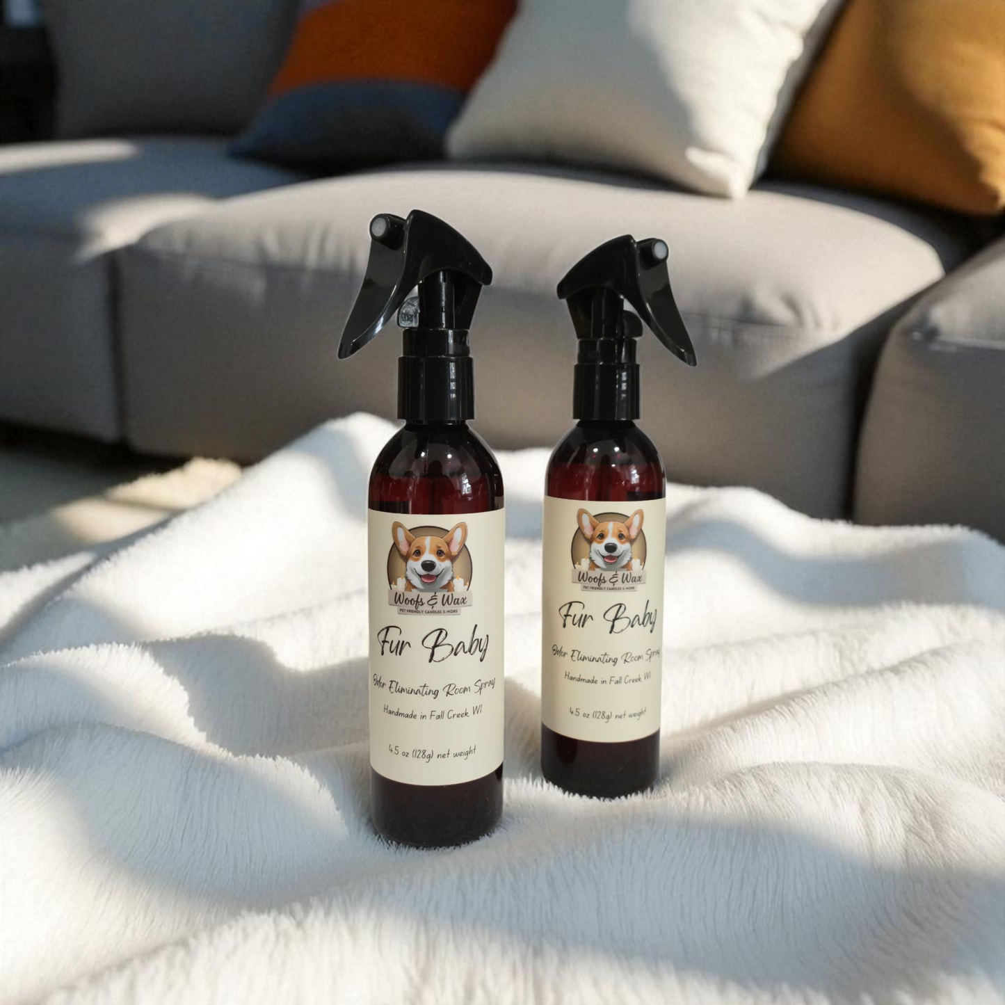 Two bottles of 'Fur Baby' pet room spray on a white blanket with a sofa in the background.