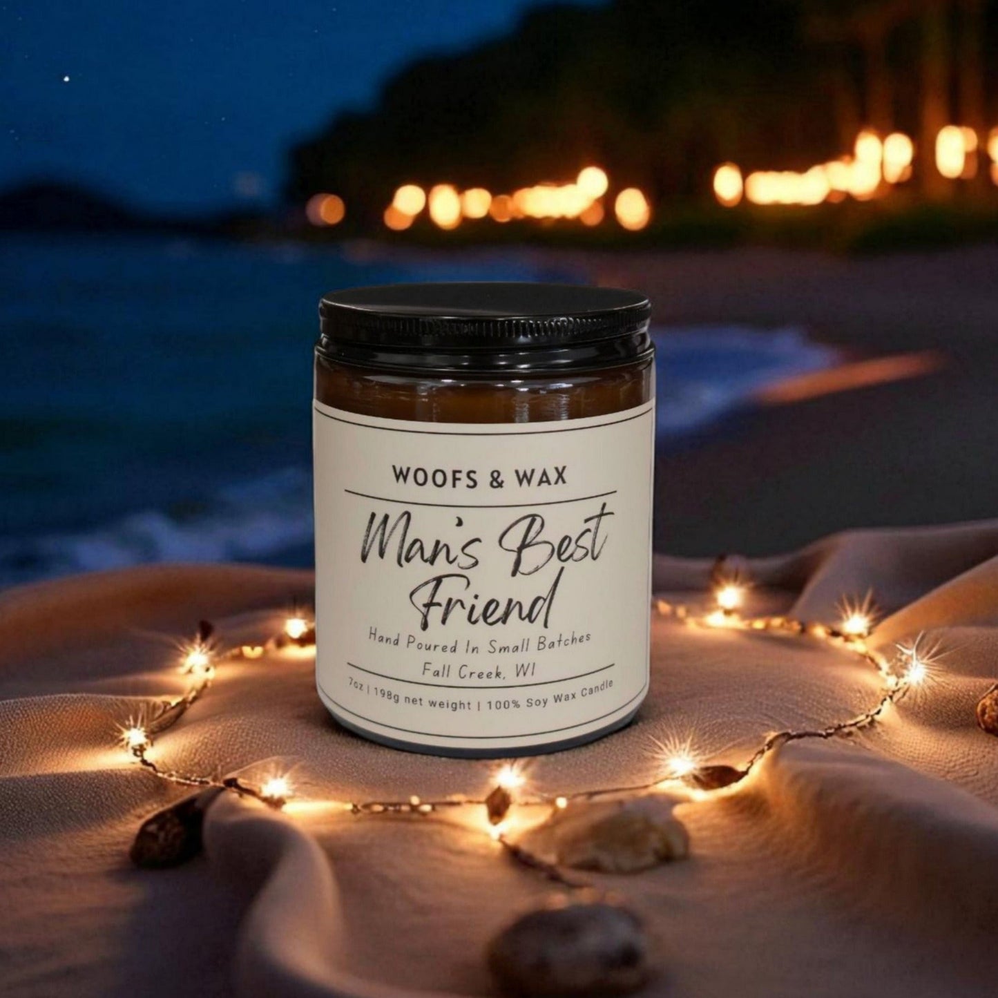 Candle labeled 'Man's Best Friend' on a beach at night with string lights and ocean view.