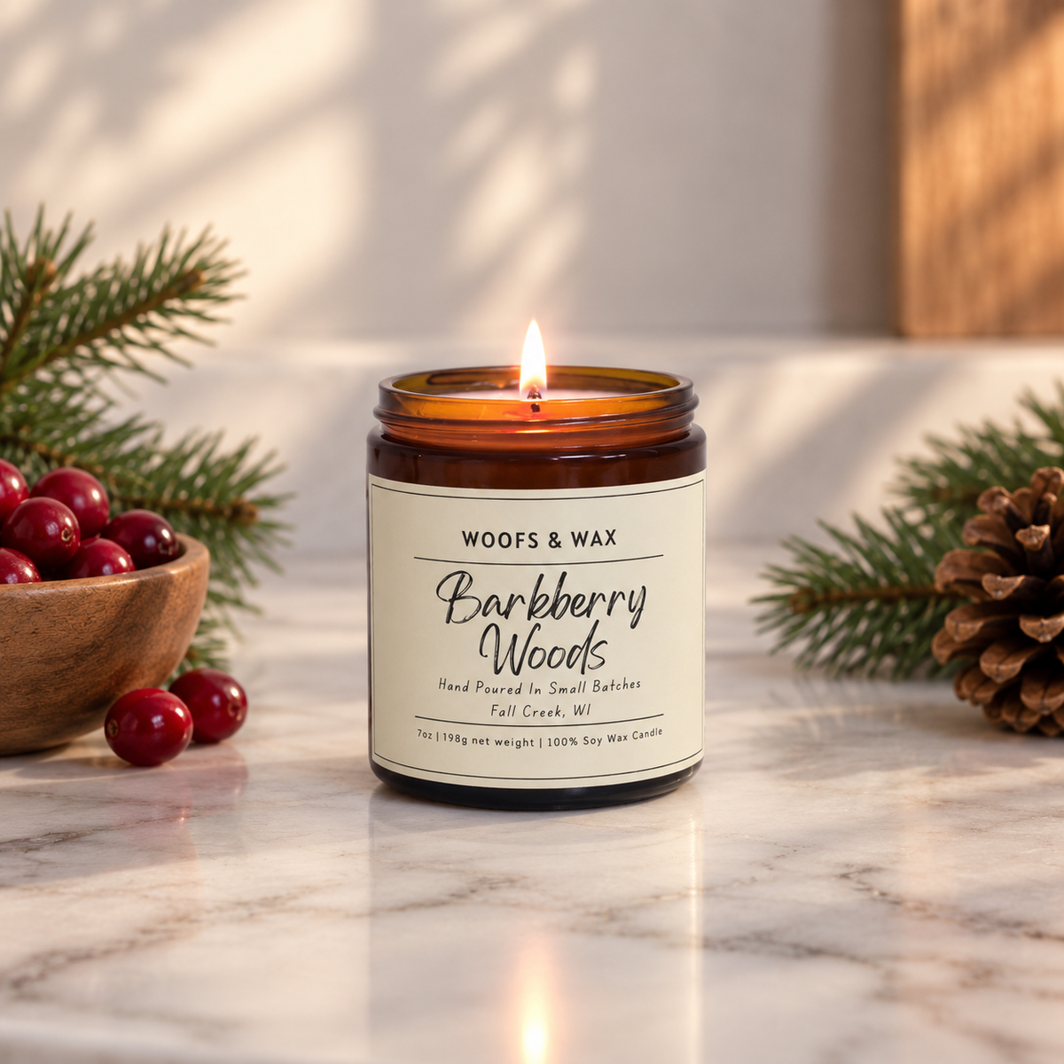 A lit amber glass jar candle with a cream label reading 'Woofs & Wax Barkberry Woods' sits on a marble surface, surrounded by pine branches, red berries, and a pinecone in a styled holiday setting.