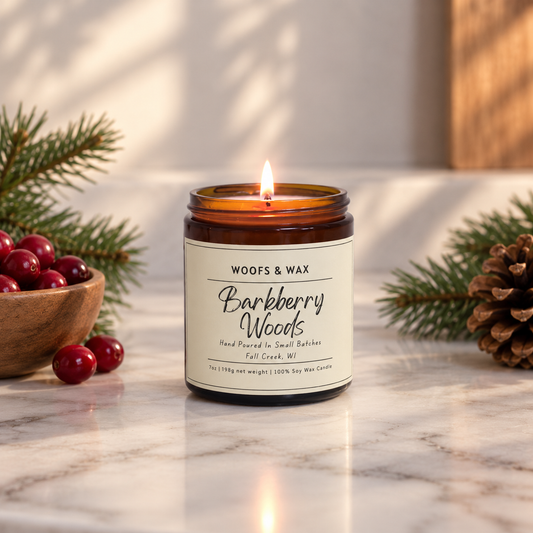 A lit amber glass jar candle with a cream label reading 'Woofs & Wax Barkberry Woods' sits on a marble surface, surrounded by pine branches, red berries, and a pinecone in a styled holiday setting.