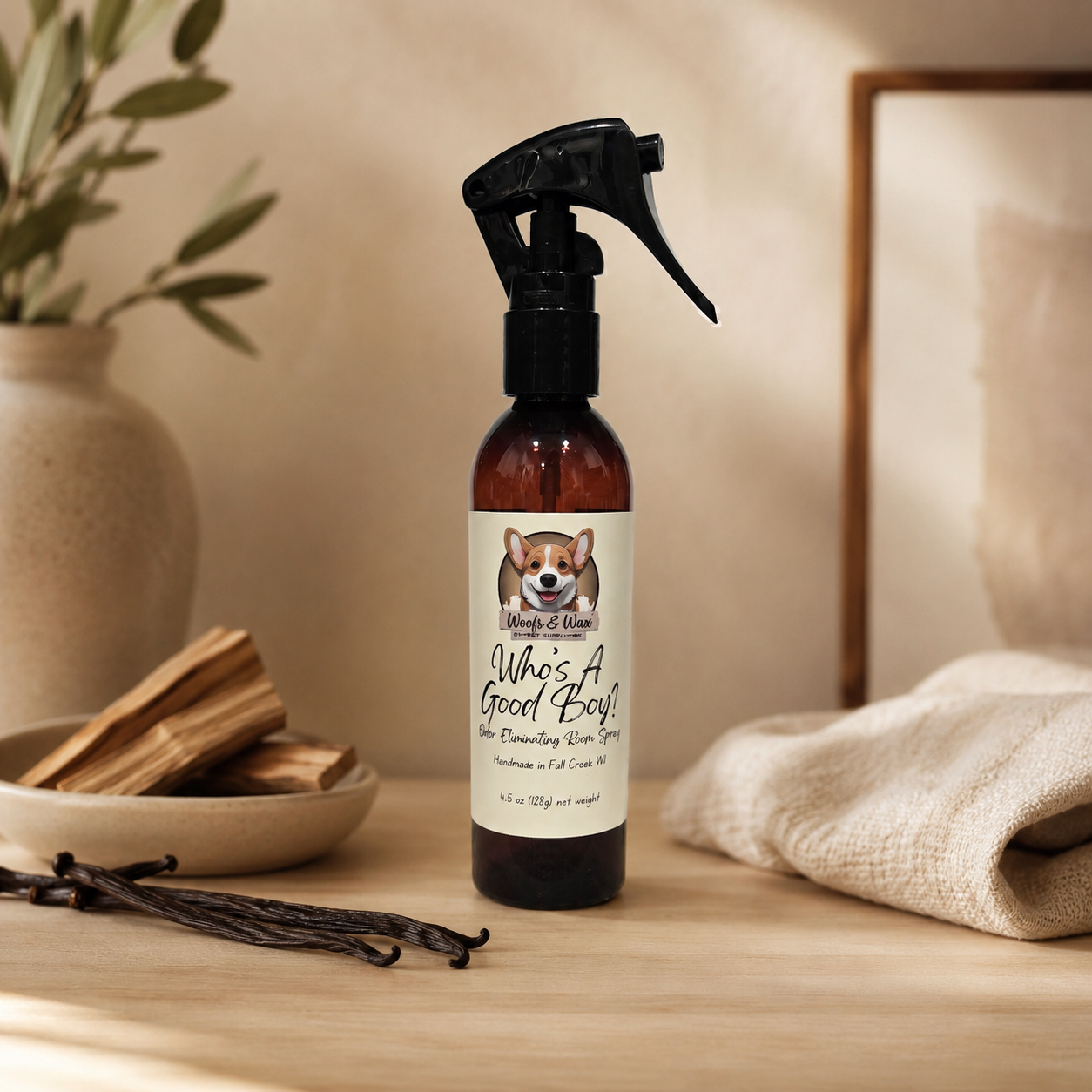 Bottle of 'Who's A Good Boy!' room spray on a wooden surface with a plant and towel in the background.