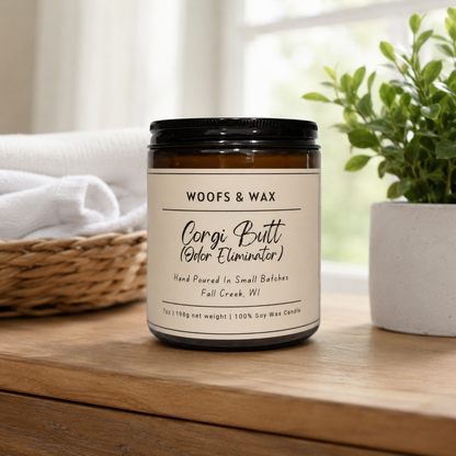 Candle labeled 'Woofs & Wax Corgi Butt Odor Eliminator' on a wooden surface with a plant and basket in the background.