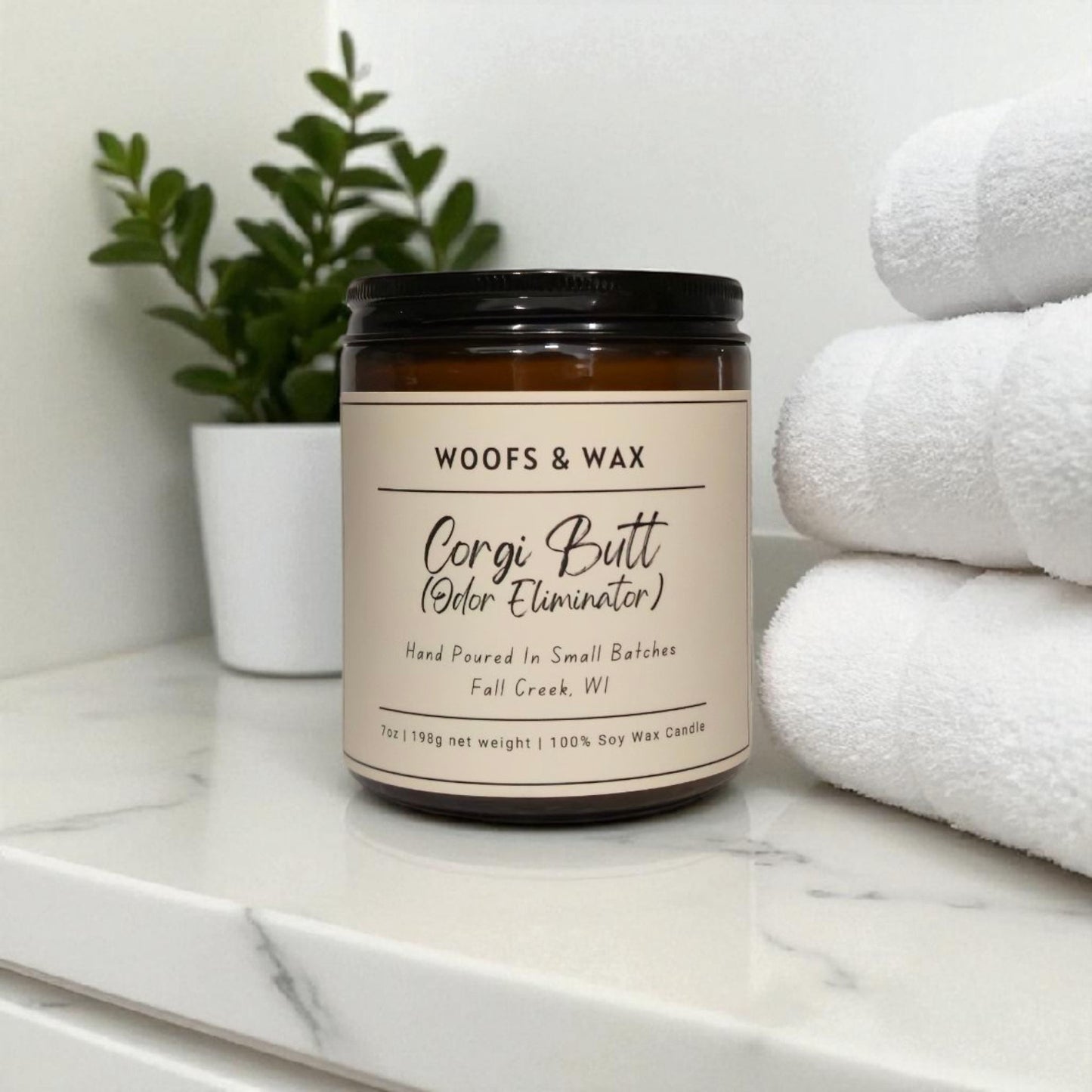 Candle labeled 'Woofs & Wax Corgi Butt Odor Eliminator' on a marble surface with towels and a plant in the background.