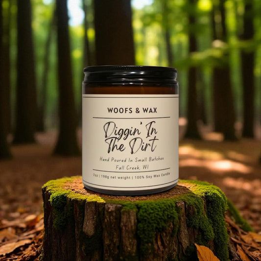Candle labeled 'Woofs & Wax Diggin' In The Dirt' on a tree stump in a forest setting