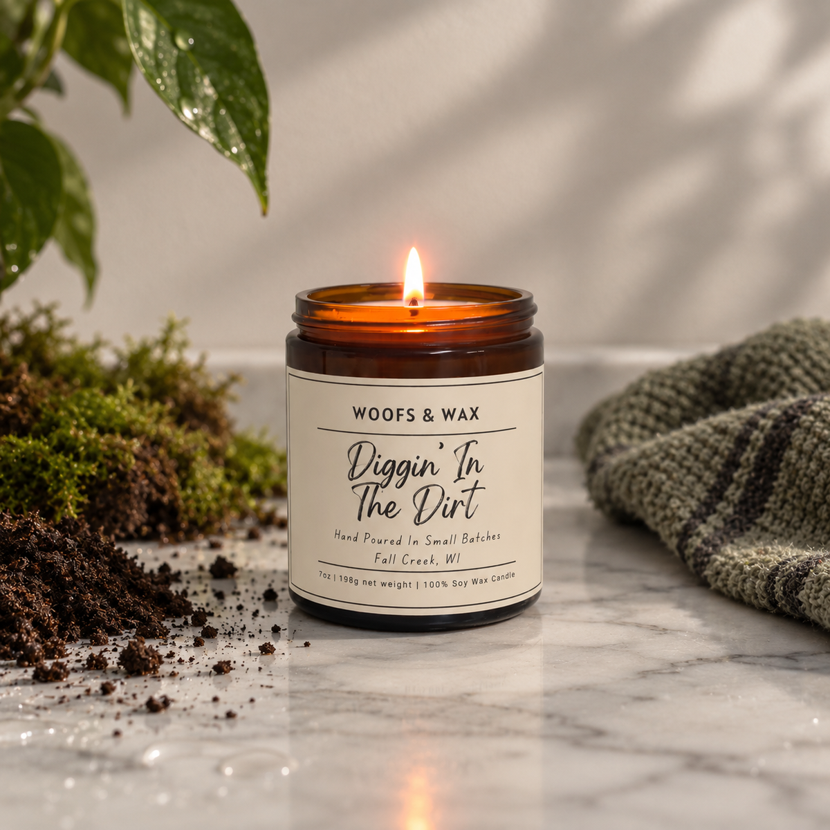 A lit amber glass jar candle with a cream label reading 'WOOF&S WAX Diggin' In The Dirt' sits on a marble surface with soil, moss, green leaves, and a knitted blanket nearby.