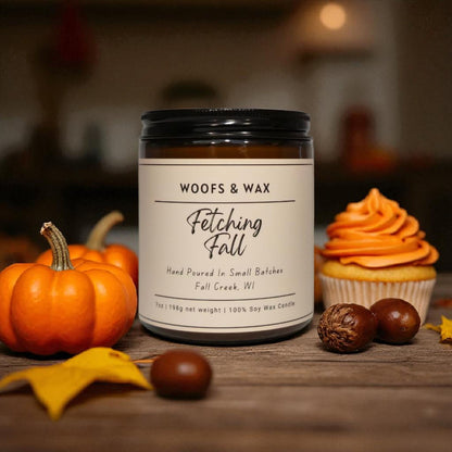 Candle labeled 'Woofs & Wax Fetching Fall' with pumpkins, a cupcake, and chestnuts on a wooden surface.