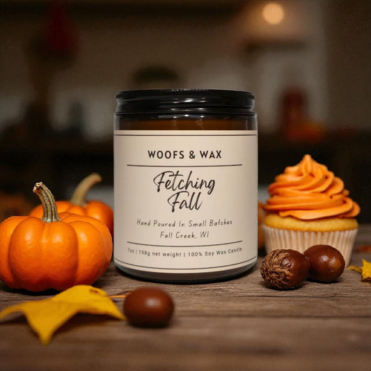 Candle labeled 'Woofs & Wax Fetching Fall' with pumpkins, a cupcake, and chestnuts on a wooden surface.