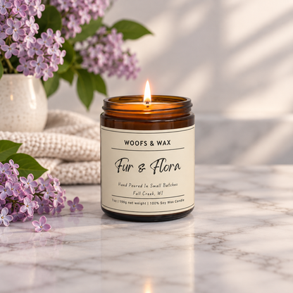A lit amber glass jar candle with a cream label reading 'WOOF&S WAX Fur & Flora' sits on a marble surface. Purple flowers and a white vase are blurred in the background, with soft natural light casting gentle shadows.