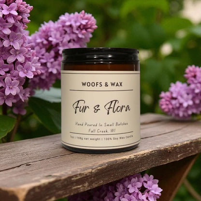 Candle labeled 'Woofs & Wax Fur & Flora' on a wooden surface with purple flowers in the background