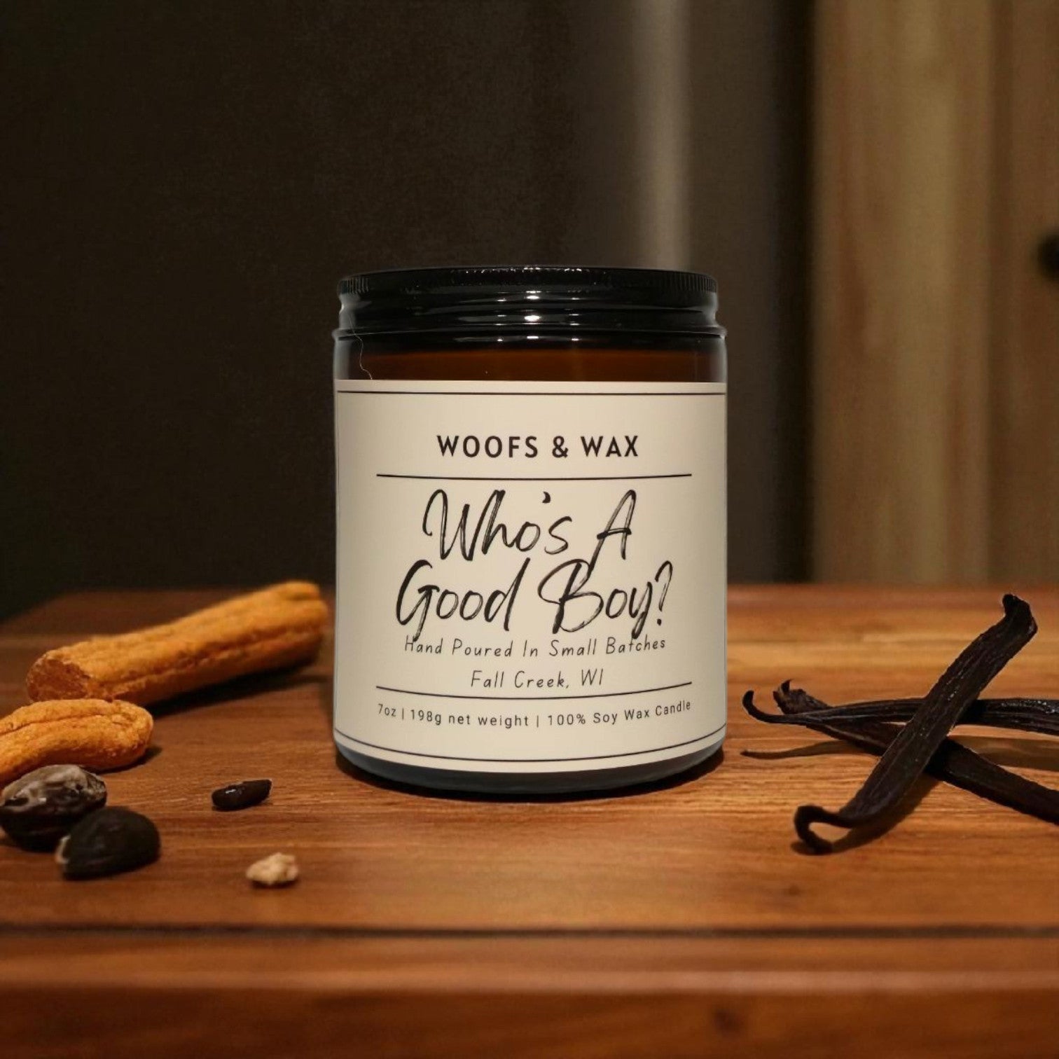 Candle jar labeled 'Woofs & Wax Whos A Good Boy?' on a wooden surface with vanilla beans and chocolate.
