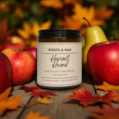 Candle jar labeled 'Woofs & Wax Harvest Hound' surrounded by apples and leaves on a wooden surface.