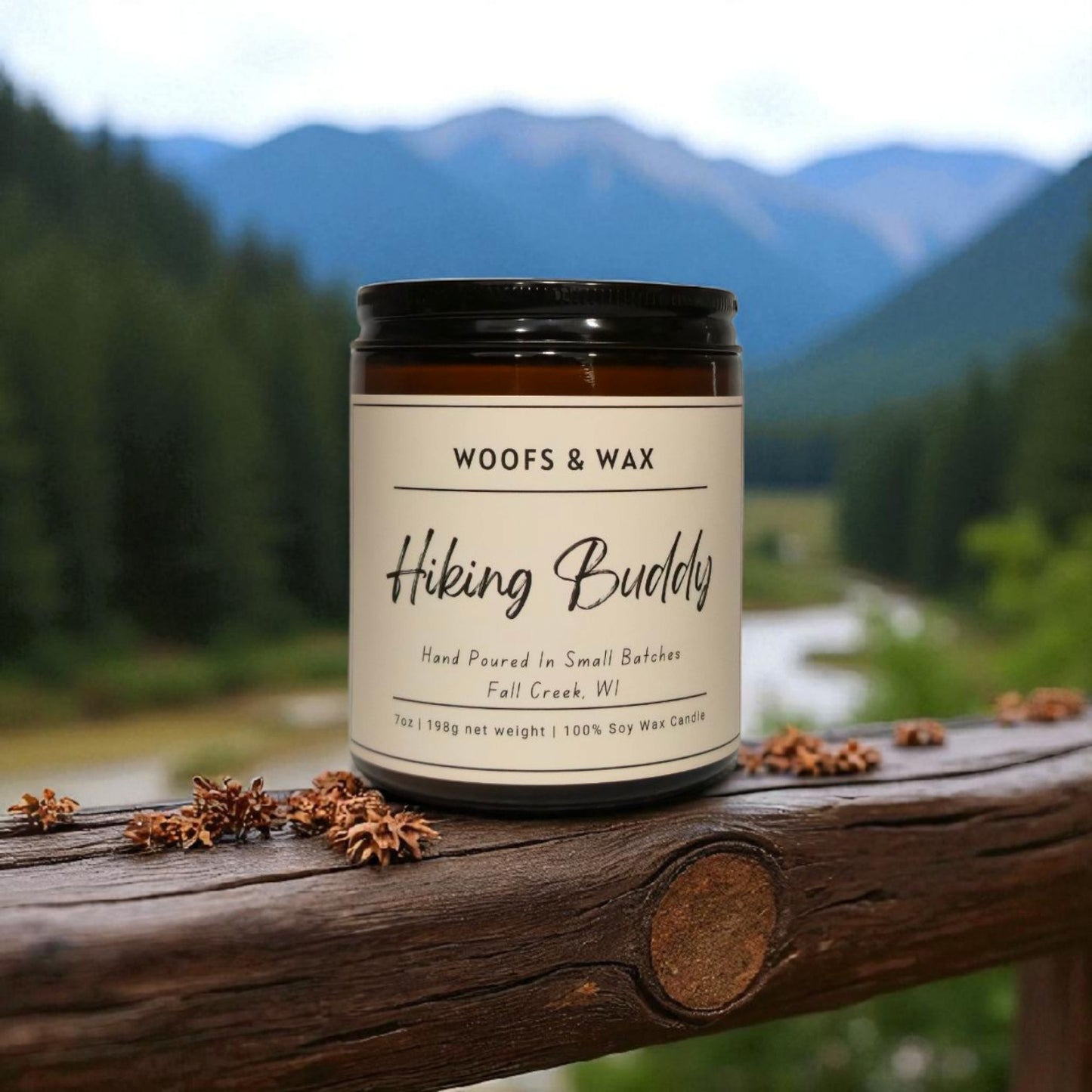 Candle labeled 'Woofs & Wax Hiking Buddy' on a wooden surface with a mountainous landscape in the background