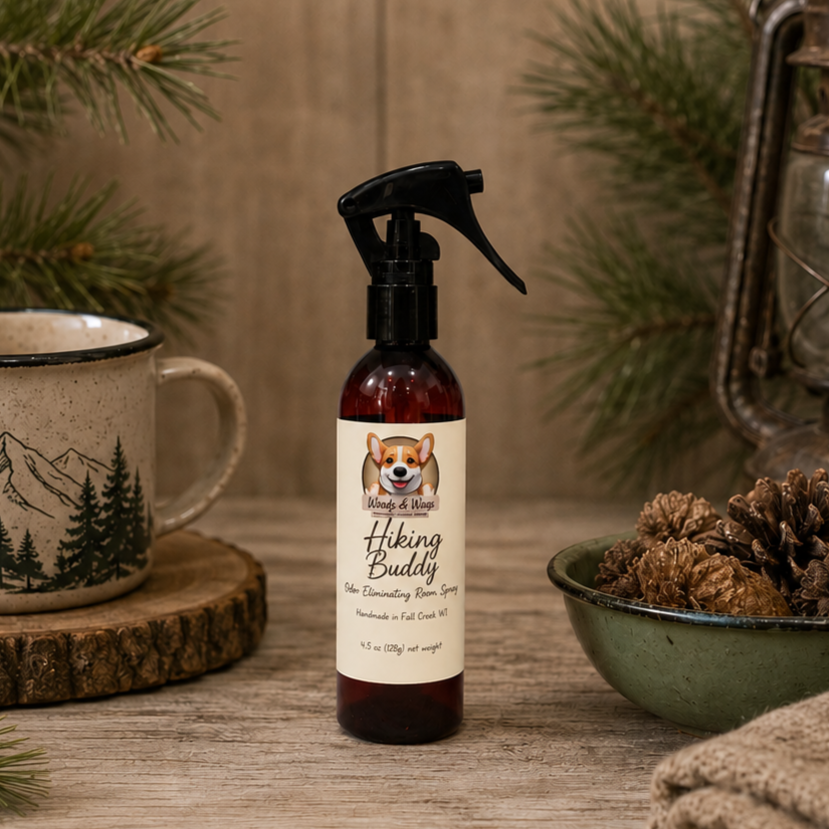 Bottle of 'Hiking Buddy' spray on a wooden surface with a mug and pine cones in the background.