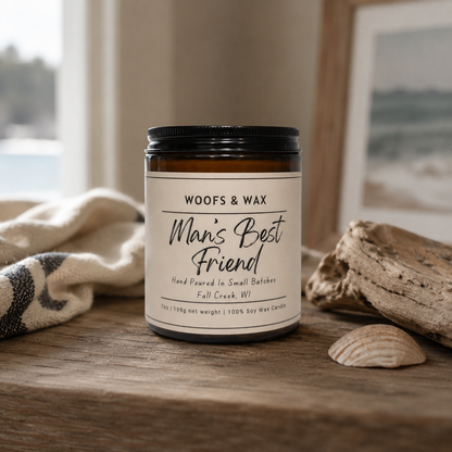 Candle jar labeled 'Man's Best Friend' by Woofs & Wax on a wooden surface with a coastal background.