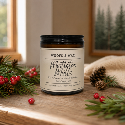 Candle labeled 'Woofs & Wax Mistletoe Mutts' on a wooden surface with holiday decorations.