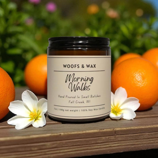 Candle labeled 'Woofs & Wax Morning Walks' surrounded by oranges and flowers on a wooden surface.