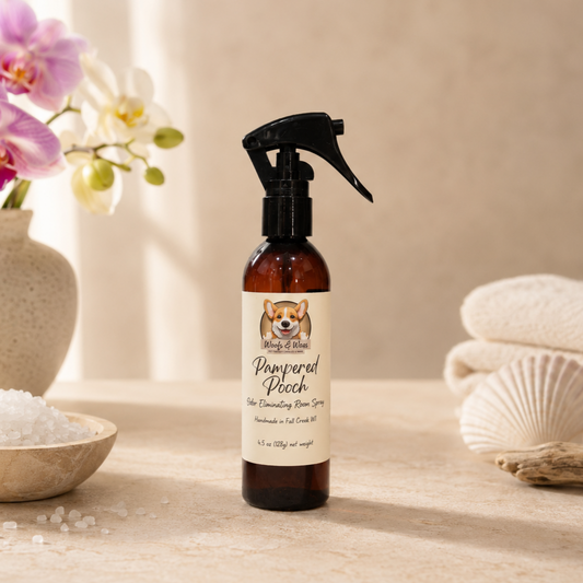 Bottle of 'Pampered Pooch' room spray on a wooden surface with flowers and towels in the background.