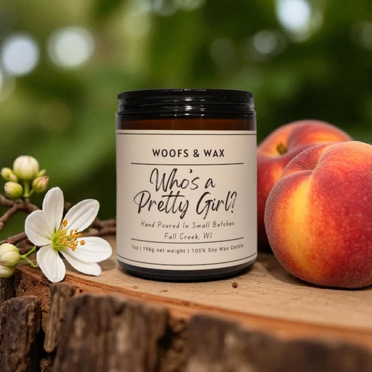 Candle jar labeled 'Woofs & Wax Who's a Pretty Girl?' with peaches and flowers on a wooden surface.