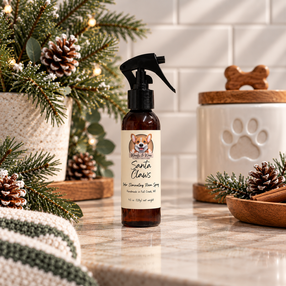 A bottle of Santa Claws room spray stands on a marble countertop in a styled kitchen scene with holiday greenery, pinecones, and a ceramic pet treat container.