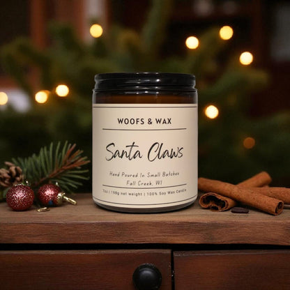 Candle labeled 'Woofs & Wax Santa Claws' on a wooden surface with festive decorations.