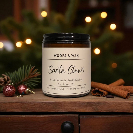 Candle labeled 'Woofs & Wax Santa Claws' on a wooden surface with festive decorations.