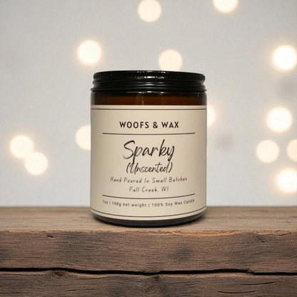 Candle jar labeled 'Woofs & Wax Sparkly Unscented' on a wooden surface with blurred lights in the background.