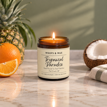 A lit amber glass jar candle labeled 'Woofs & Wax Tropawcal Paradise' sits on a marble surface with pineapple, orange slices, a coconut half, and a folded towel in the background.