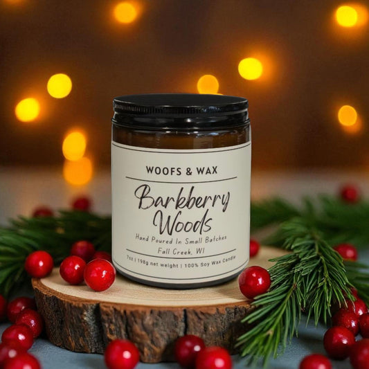 Candle jar labeled 'Woofs & Wax Barkberry Woods' on a wooden stump with berries and greenery, blurred lights in the background.