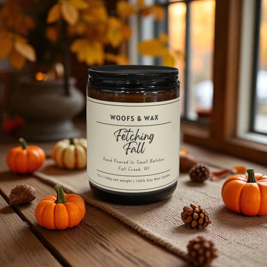 Candle labeled 'Woofs & Wax Fetching Fall' on a wooden surface with pumpkins and pinecones.