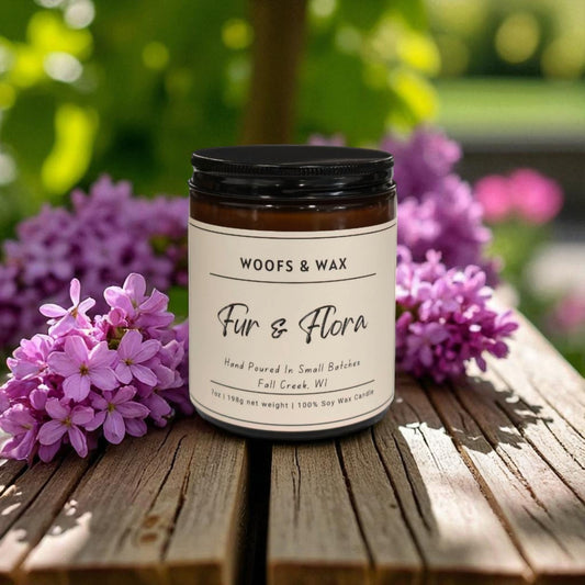Jar of 'Fur & Flora' candle by Woofs & Wax on a wooden surface with purple flowers in the background