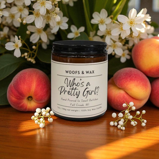 Candle labeled 'Who's a Pretty Girl?' by Woofs & Wax with peaches and flowers on a wooden surface