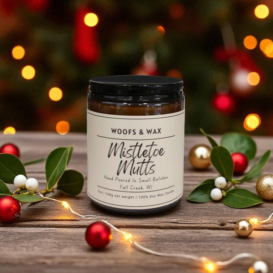 Candle jar labeled 'Woofs & Wax Mistletoe Mutts' on a wooden surface with Christmas decorations.