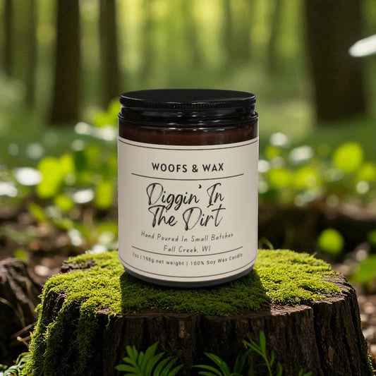 Candle jar labeled 'Woofs & Wax Diggin' In The Dirt' on a mossy log with a forest background