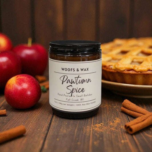 Jar of 'Pawtum Spice' candle from Woofs & Wax with apples, cinnamon sticks, and a pie in the background on a wooden surface.