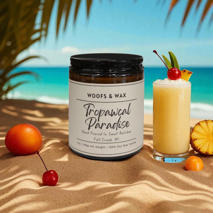 Candle labeled 'Tropical Paradise' by Woof & Wax on a sandy beach with a cocktail and fruit.