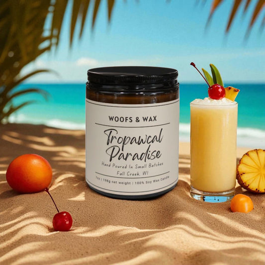 Candle labeled 'Tropical Paradise' by Woof & Wax on a sandy beach with a cocktail and fruit.
